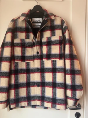 Rutig overshirt från Essentiel Antwerp - Säljer en oversized rutig overshirt från Essentiel Antwerp. Jackan har stora bröstfickor med knappar, klassisk krage och stängs med knappar framtill. Färgerna är beige, svart, grå och röd. Tillverkad i ullblandning, perfekt för lager på lager under höst och vinter.