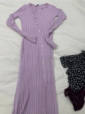 Långärmad lila ribbad klänning - Superfin långärmad lila klänning med ribbad struktur och knappar hela vägen framtill. Klänningen är lång och har en v-ringad halsringning. Perfekt för dig som gillar en bekväm och snygg stil med lite extra edge.