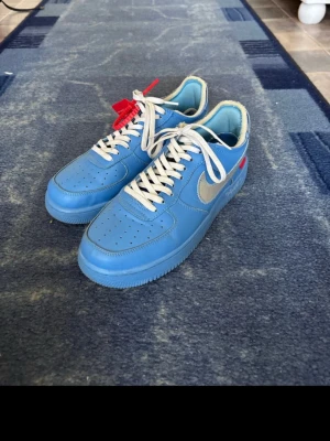 Nike Air Force 1 Off-White  - Säljer ett par Nike Air Force 1 Off-White i blått med silver swoosh och vit text på sidan. Då jag inte använder dom längre. Skorna har vita snören, röd tag och är tillverkade i skinn. Klassisk perforerad tå och platt sula. Perfekta för dig som gillar streetwear och unika sneakers.