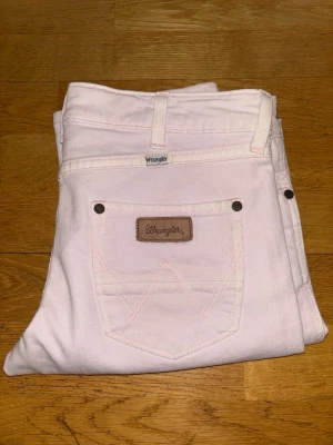 Ljusrosa jeans från Wrangler - Säljer ett par ljusrosa jeans från Wrangler med klassisk femficksdesign och raka ben. Byxorna har snygga detaljer med nitar vid fickorna och Wrangler-logga på bakfickan. Materialet är 60% bomull och 40% polyester för skön komfort.