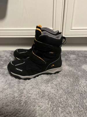 Svarta Viking vinterstövlar strl 39 - Säljer ett par svarta Viking Bluster II GTX vinterstövlar med grå och gula detaljer. Skorna har grov sula med gult mönster undertill, två kardborreband och är fodrade med grått tyg. Perfekta för kalla dagar och snöiga äventyr. Nypris 1399kr säljs för 699kr pris kan diskuteras!