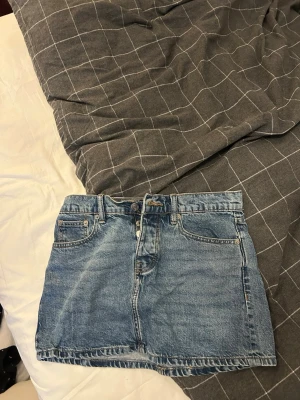 Blå jeanskjol från 157, storlek XS - Säljer en klassisk blå jeanskjol från 157 i modellen Maddy. Kjolen har fem fickor, bälteshällor och stängs med knapp och dragkedja framtill. Perfekt basic-plagg att styla med allt från hoodies till crop tops.