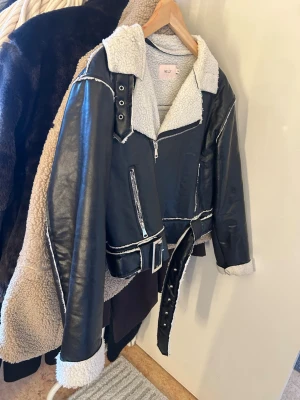 Svart teddyjacka med skinn från Nelly - Svart teddyjacka från Nelly med vita detaljer och fuskpälsfoder. Jackan har bred krage, dragkedjor, bälte i midjan och coola sömmar. Materialet är skinnimitation och teddy, perfekt för dig som vill ha en edgy look med extra värme.