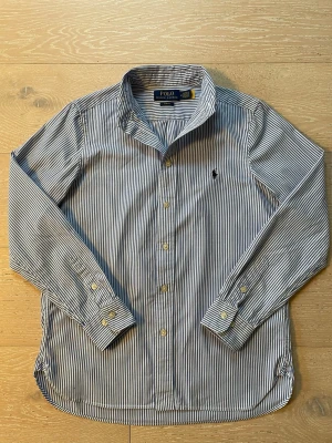 Randig skjorta från Polo Ralph Lauren - Säljer en blåvit randig skjorta från Polo Ralph Lauren i slim fit. Skjortan har klassisk krage, långa ärmar med manschett och knappar framtill. Broderad logga på bröstet och tillverkad i mjuk bomull. Perfekt för dig som gillar stilrena och tidlösa plagg. Mycket bra skick, inga defekter. Fråga gärna om ni undrar något! Prisförslag uppskattas