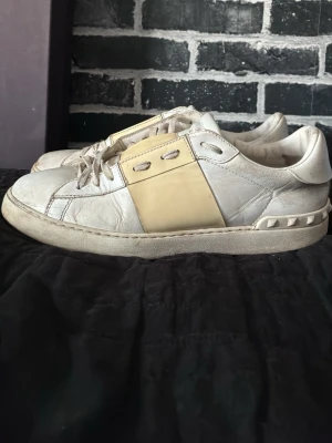 Valentino Garavani Rockstud sneakers - Säljer ett par vita Valentino Garavani Rockstud sneakers i skinn med ikoniska nitar runt hälen och en beige detalj på sidan. Skorna har snörning och en chunky sula för en cool streetwear-look. Perfekta för dig som vill ha något unikt och lyxigt i garderoben.