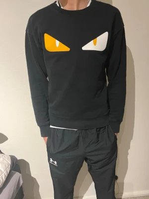 Svart Fendi sweatshirt med ögon - Säljer en svart sweatshirt från Fendi med ikoniska gula och vita ögon framtill och Fendi-logga i gult bak i nacken. Tröjan har rund hals, långa ärmar och är gjord i mjuk bomull. Perfekt för dig som vill sticka ut med en unik designertröja. Pris kan diskuteras och hör av er om ev frågor. 