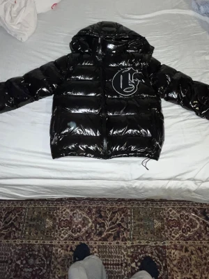 Svart Corvidae v2 shiny jacka - Säljer en glansig svart pufferjacka från Corvidae med avtagbara ärmar så du kan bära den som väst. Jackan har huva, stor logga på bröstet och dragkedja framtill. Storleken är en SX men sitter mer som en S eller M