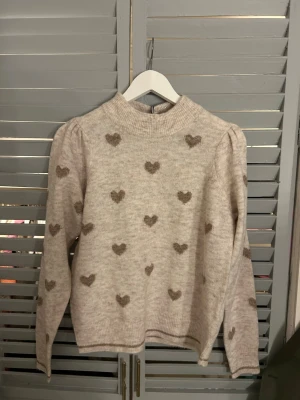 Beige stickad tröja med hjärtan Freequent - Superfin beige stickad tröja från Freequent med bruna hjärtan över hela. Tröjan har högre ribbad hals, lång ärm och en lite loose passform. Perfekt till jeans eller kjol för en söt och mysig look.