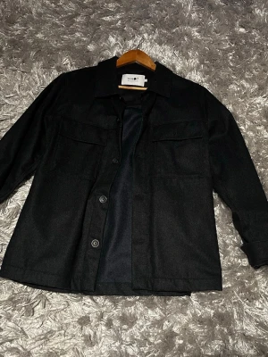 Svart overshirt från NN07 - Säljer en svart overshirt från NN07. Helt nyköpt från Nordiska Kompaniet. Medklassisk krage och stora bröstfickor. Jackan har knäppning framtill med fyra knappar och är tillverkad i ett mjukt material. 