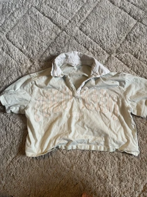 Ljusbeige henley från H&M - Kortärmad ljusbeige henley från H&M med vit krage och trycket 'COLORADO' framtill. Skjortan har en relaxed, croppad passform och knappar vid halsen. Perfekt för en chill och avslappnad stil.