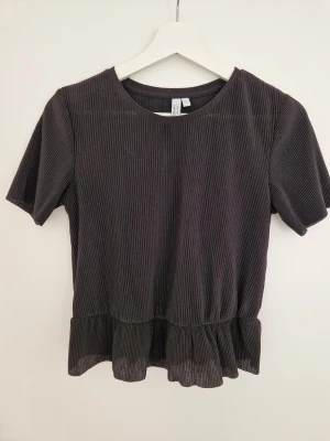 Svart ribbad topp från & Other Stories - Säljer en svart ribbad topp från & Other Stories med rund halsringning och korta ärmar. Toppen har volangkant nedtill som ger en söt touch och är tillverkad i ett mjukt, stretchigt material. Perfekt att styla med jeans eller kjol. Väl använd, lite nopprigt på tyget.
