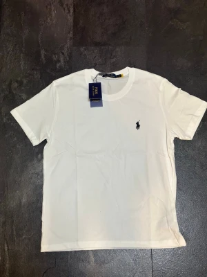 Vit t-shirt från Polo Ralph Lauren - Klassisk vit t-shirt från Polo Ralph Lauren med rund hals och korta ärmar. Diskret broderad logga i svart på bröstet. Tillverkad i mjuk bomull för en clean och enkel look. Perfekt basplagg till alla outfits.