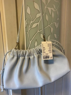 Ljusblå axelväska - Säljer en ljusblå axelväska i syntetmaterial med trendig snörning upptill. Väskan har en avtagbar axelrem och en mjuk, puffig form som ger en modern look. Perfekt för dig som vill ha en stilren och enkel accessoar.