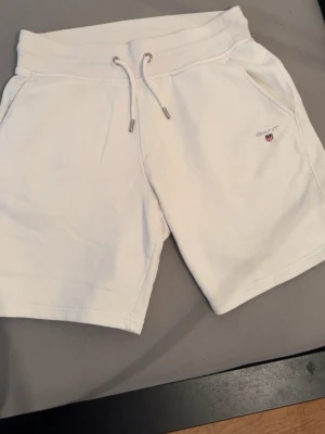 Vita sweatshorts från GANT - Säljer ett par vita sweatshorts från GANT med dragsko i midjan och broderad logga på ena benet. Shortsen har två sidofickor fram och en bakficka. Tillverkade i mjuk bomull, perfekta för chill dagar eller sommarhäng.