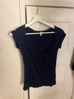 Mörkblå topp med spetskant från Nelly - Säljer en mörkblå topp från Nelly i storlek S. Toppen har korta ärmar, rundad urringning och en söt spetskant längs halsringningen. Tillverkad i mjukt material som känns skönt mot huden. Perfekt att styla med jeans eller kjol.