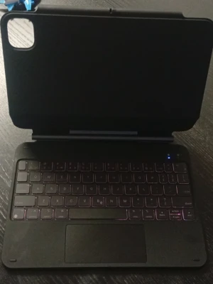 iPad tangentbord med fodral - Snyggt och modernt tangentbord med fodral för iPad. Utrustad med bakgrundsbelysta tangenter och stor styrplatta. Fodralet har utskärning för kamera och skyddar din iPad effektivt. Tangentbordet verkar vara i gott skick utan synligt slitage.