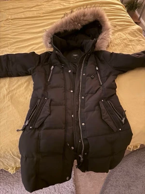 Svart vinterjacka - Säljer en svart lång dunjacka från Rockandblue med stor huva och fluffig fuskpäls. Jackan har flera dragkedjeförsedda fickor, snygga detaljer och patch på ärmen. Perfekt för kalla dagar och riktigt varm. Dragsko i midjan för bästa passform.