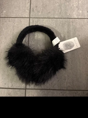 Svart fluffig öronmuff  från JYOTHI - Säljer en svart öronmuffar från JYOTHI med superfluffig och mjuk känsla och är perfekt för att addera lite extra stil till din outfit. Materialet är pälsimitation har en klassisk passform.