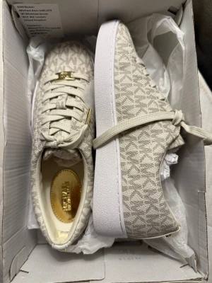 Beige sneakers från Michael Kors - Snygga beige sneakers från Michael Kors med monogrammönster och guldfärgade detaljer på snörningen. Skorna har rund tå, platt vit sula och är tillverkade i textil med syntetdetaljer. Perfekta för dig som gillar stilrena och trendiga sneakers. Så fina till inför våren💕