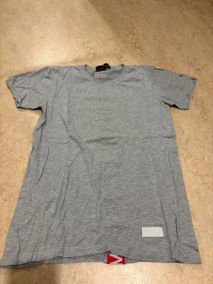 Grå t-shirt från Concrete Humanity - Snygg grå t-shirt från Concrete Humanity i mjuk bomull. På baksidan finns en bred röd rand med vit text 'CONCRETE HUMANITY' som ger en unik look. Klassisk passform och rund halsringning. Perfekt till jeans eller shorts.