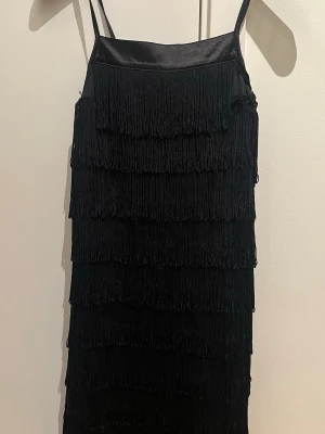 Svart fransklänning från Vero Moda - Säljer en svart kort klänning från Vero Moda med tunna axelband och flera lager fransar som ger en riktigt cool look. Perfekt för dig som vill sticka ut och älskar detaljer. Klänningen är i mjukt syntetmaterial och har en snygg, rak passform.