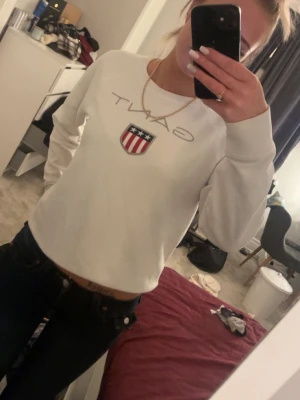 Vit sweatshirt från GANT med emblem - Säljer en fräsch vit sweatshirt från GANT med stort tryckt logotyp och ett amerikanskinspirerat emblem på bröstet. Tröjan har rund hals, långa ärmar och klassisk passform. Perfekt till jeans för en clean och sportig look.