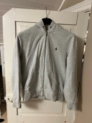 Grå Ralph Lauren Hoodie M - En grå Ralph lauren hoodie medium. Den är i nyskick och passar bra på både tjejer och killar. Perfekt för kyliga dagar som nu. Tyvärr medföljer inga snören.
