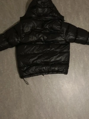 Svart pufferjacka från Polo Ralph Lauren - Säljer en svart pufferjacka från Polo Ralph Lauren med huva och dragkedja. Jackan har en klassisk röd logga på bröstet, stora vadderade partier och snörning nertill. Perfekt för kalla dagar och riktigt snygg till streetwear-stilen.