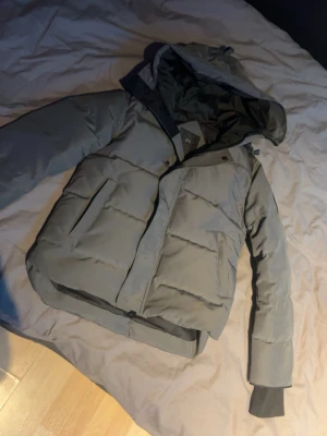 Grå pufferjacka från Canada Goose - Säljer en grå pufferjacka från Canada Goose i storlek S. Jackan har huva, stora fickor framtill och dragkedja med tryckknappar. Perfekt för kalla dagar och riktigt skön att bära. Klassisk design med diskret logga på dragkedjan.