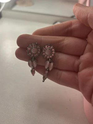 Rosa vintage-inspirerade örhängen - Säljer ett par unika örhängen med rosa stenar och pärlliknande detaljer. Örhängena har en rund topp med gnistrande rosa stenar runt en större mittsten och hängande droppformade dekorationer. Perfekt accessoar för dig som gillar retrostil och vill sticka ut.
