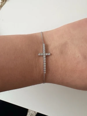 Smyckad silverarmband med kors - Tunt armband i silverfärg med ett gnistrande kors dekorerat med små stenar. Stilren och enkel kedja som ger ett elegant intryck. Perfekt accessoar för dig som gillar diskreta men snygga detaljer.