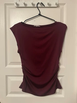 Vinröd topp från H&M - Säljer en vinröd topp från H&M i mjukt och stretchigt material. Toppen har en enkel, ärmlös design med lätt rynkade sidor som ger en snygg siluett. Perfekt att styla med jeans!💓😽