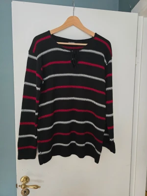 Randig stickad tröja från Elitzia - Säljer en svart stickad tröja från Elitzia med vita och röda horisontella ränder. Tröjan har lång ärm, rund halsringning med snörning och en relaxed fit. Perfekt för dig som gillar en enkel men ändå snygg look.