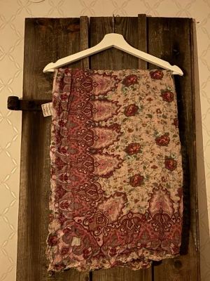 Blommig sjal från Odd Molly - Unik sjal från Odd Molly i tunn bomull med blommigt mönster i rött, grönt och beige. Sjalen har ett bohemiskt tryck med rosor och dekorativa detaljer i vinröda toner. Perfekt för dig som vill sticka ut med en färgstark och mönstrad look. Storlek; ca. 1 * 1m