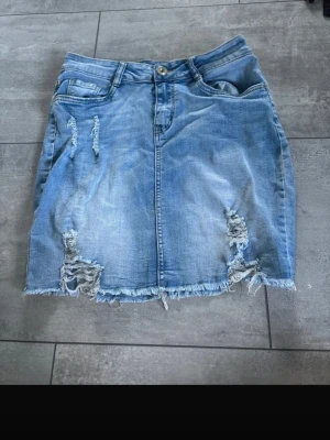 Blå jeanskjol med slitningar - Säljer en blå jeanskjol med rå kant och slitna detaljer både fram och bak. Kjolen har klassiska fickor, bälteshällor och en knapp framtill. Perfekt för dig som gillar en avslappnad och trendig look.