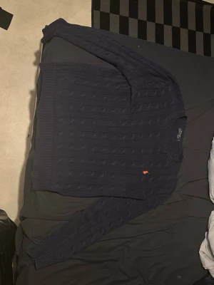 Mörkblå kabelstickad tröja Polo Ralph Lauren - Klassisk mörkblå kabelstickad tröja från Polo Ralph Lauren med orange broderad logga på bröstet. Tröjan har rund halsringning, ribbade muddar och är långärmad. Perfekt för dig som gillar stilrena och tidlösa plagg. PRIS EJ HUGGET I STEN