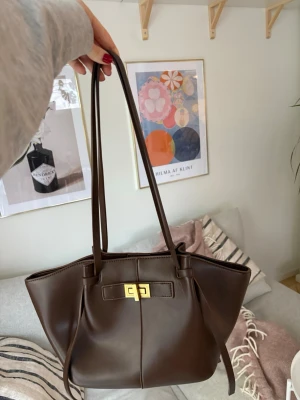 Mörkbrun axelväska med gulddetalj - Säljer en stilren mörkbrun axelväska i skinnimitation med långa axelband och snygg guldfärgad vridlåsdetalj framtill. Väskan har en rymlig form och minimalistisk design som passar perfekt till vardagslooken. Perfekt för dig som vill ha plats för allt du behöver under dagen.