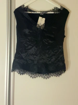 Svart satinlinne med spets Ever Since 1973 - Supersnygg svart linnetopp från Ever Since 1973 i glansigt satinmaterial. Dekorerad med spetsdetaljer längs halsringning och nederkant för en extra elegant touch. Ärmlös och med v-ringad skärning. Med orginal prislappen på