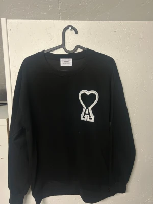 Ami pari - Svart sweatshirt från Ami Alexandre Mattiussi med stor vit och svart hjärtdetalj och bokstaven A på bröstet. Klassisk rund halsringning och långa ärmar. Tillverkad i mjuk bomull för en skön och avslappnad look. Den är knappat använd S/xs