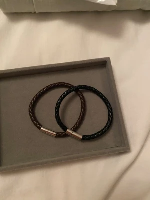 2 Riktigt snygga armband  - Bilderna visar två flätade armband i svart och brunt läder med metallås, Helt nya.
