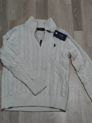 Riktig och helt ny Vit stickad tröja Polo Ralph Lauren - Stickad vit tröja från Polo Ralph Lauren i storlek S. Tröjan har hög krage, half zip-dragkedja med läderdetalj och klassisk marinblå logga broderad på bröstet. Ribbstickade muddar och nederkant ger en snygg finish. Perfekt för kyliga dagar.