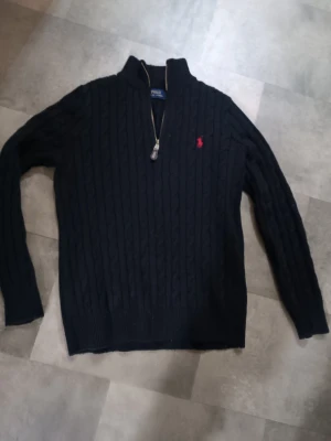 Svart stickad tröja från Polo Ralph Lauren - Svart kabelstickad tröja från Polo Ralph Lauren med halv dragkedja och röd broderad logga på bröstet. Tröjan har lång ärm, ribbad krage och muddar. Perfekt för dig som gillar klassisk stil med en modern touch.