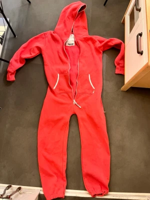 Röd jumpsuit från Ziprall med huva - Mysig röd jumpsuit från Ziprall med vit dragkedja hela vägen och två fickor framtill. Har en stor huva och muddar vid ärm- och benslut. Perfekt för chill dagar hemma eller när du vill vara extra bekväm.
