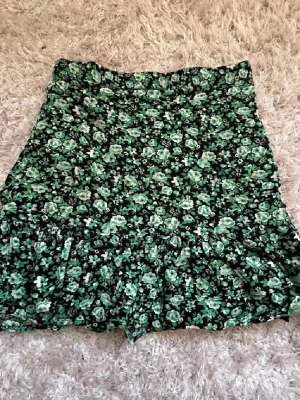 Grön blommig volangkjol - Supersöt kort kjol med grönt och vitt blommönster på svart botten. Kjolen har volangkant nedtill som ger en extra touch och flowy känsla. Perfekt för dig som vill ha en lekfull och trendig stil.