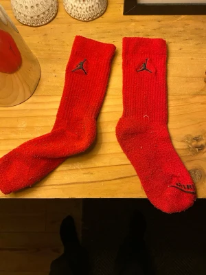 Röda Jordan strumpor med logga - Säljer ett par röda Jordan strumpor med svart logga och texten 'JORDAN' vid tårna. Strumporna är ribbade upptill och har en sportig look. Perfekta för dig som gillar streetwear och vill ha något som sticker ut. Hör av dig