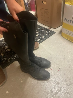 Svarta ridstövlar från CRW - Säljer ett par höga svarta ridstövlar från CRW i skinn med snörning framtill och dragkedja längs baksidan. Klassisk design med rund tå och diskreta detaljer. Perfekta för dig som vill ha stilrena och funktionella stövlar till ridning.