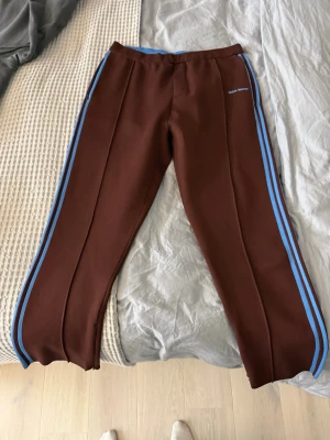 Adidas x Wales Bonner bruna mjukisbyxor - Bruna mjukisbyxor från Adidas och Wales Bonner med ljusblå ränder längs sidorna och broderad logga. Byxorna har resår och snörning i midjan samt raka ben. Tillverkade i ett mjukt syntetmaterial som känns skönt mot huden. Perfekta för en chill och sportig look.