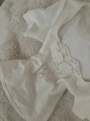 Vit bolerotopp med spetsdetalj - Supersöt vit bolerotopp med korta ärmar och bred, fyrkantig urringning. Framtill finns en dekorativ spetsdetalj med blommönster som ger en romantisk vibe. Materialet känns mjukt och stretchigt, perfekt till sommarens outfits.