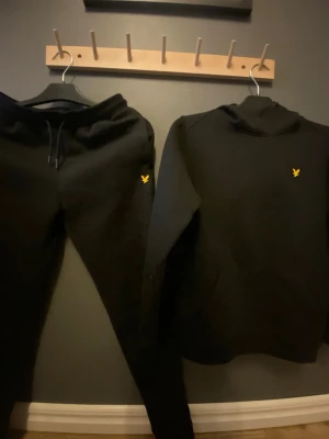 Svart Lyle & Scott huvtröja - Säljer en svart huvtröja från Lyle & Scott med den klassiska gula fågeln broderad på bröstet. Tröjan har huva och dragsko, enkel och stilren design som passar till det mesta. Perfekt för dig som gillar streetwear och vill ha något bekvämt. Hör av dig! Den är lite knottrig men inte mycket.