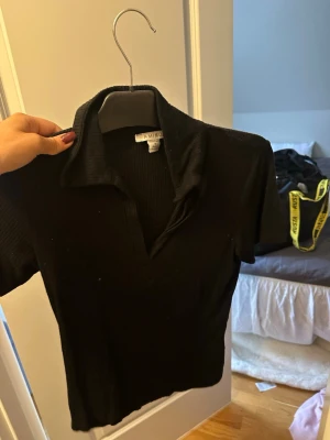 Svart ribbad topp från Amisu - Säljer en svart ribbad topp från Amisu med krage och v-ringad öppning framtill. Toppen har korta ärmar och är i stretchigt material som sitter snyggt på kroppen. Perfekt till jeans eller kjol för en clean look.